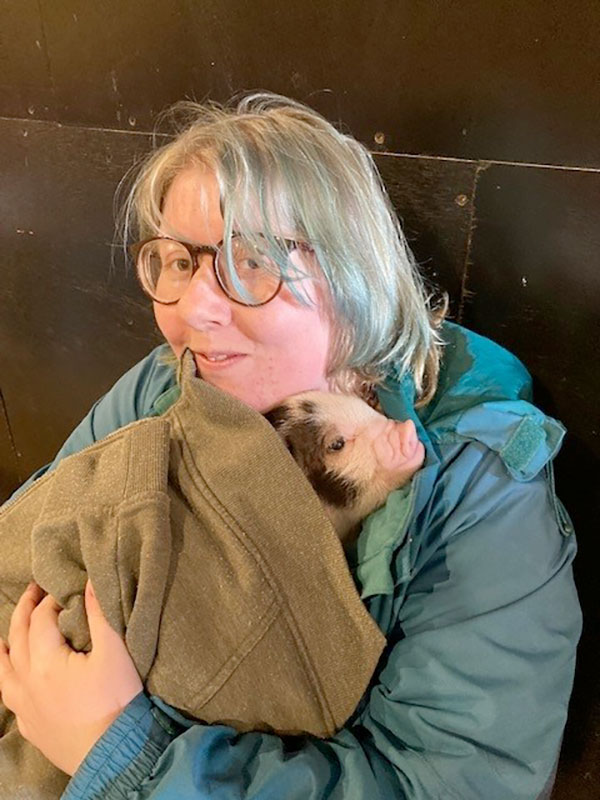 Photo of a smiling lady cuddling a piglet in a blanket.